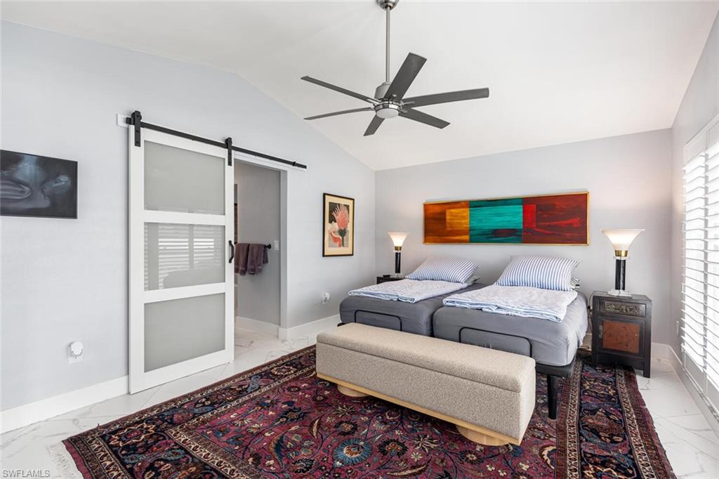 Bedroom showing porcelain tile flooring, a barn door, ceiling fan, and lofted ceiling.  Windows have wooden plantation shutters.