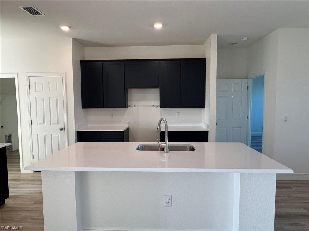 Kitchen featuring a sink, a center island with sink, light wood-type flooring, dark cabinetry, and recessed lighting