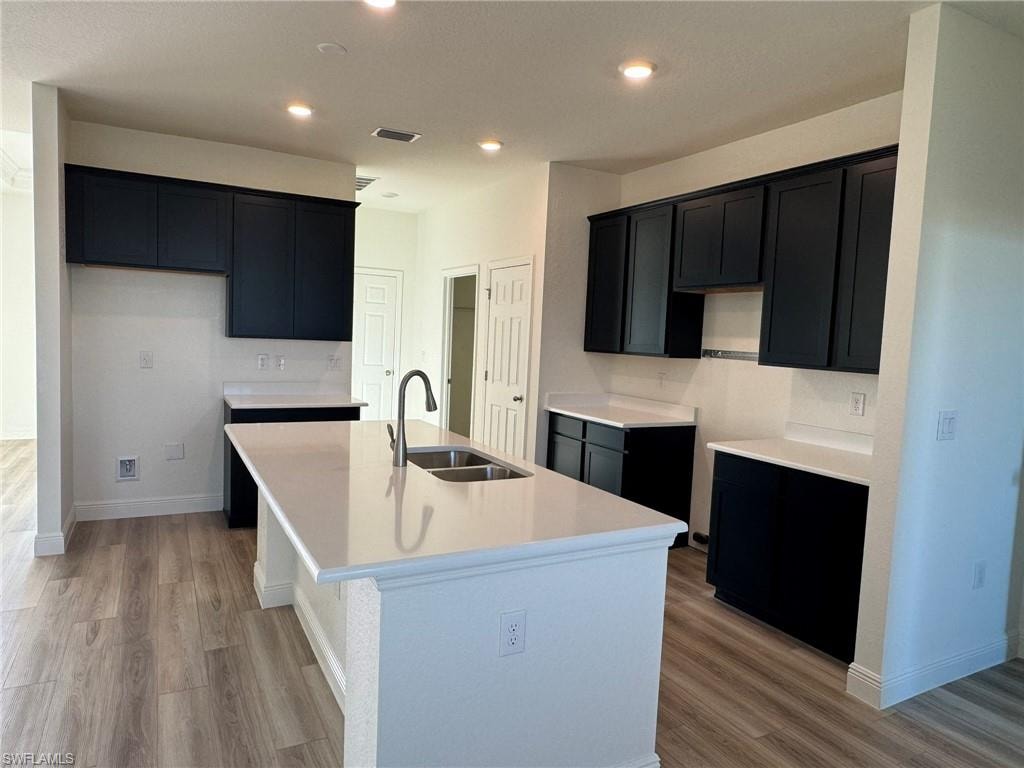 Kitchen featuring a sink, an island with sink, light wood-style floors, dark cabinets, and recessed lighting