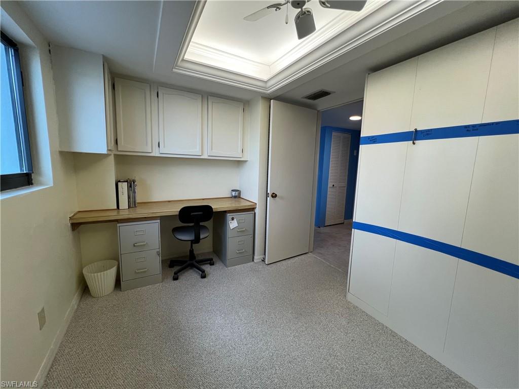 Unfurnished office with light colored carpet, ceiling fan, a raised ceiling, and built in desk
