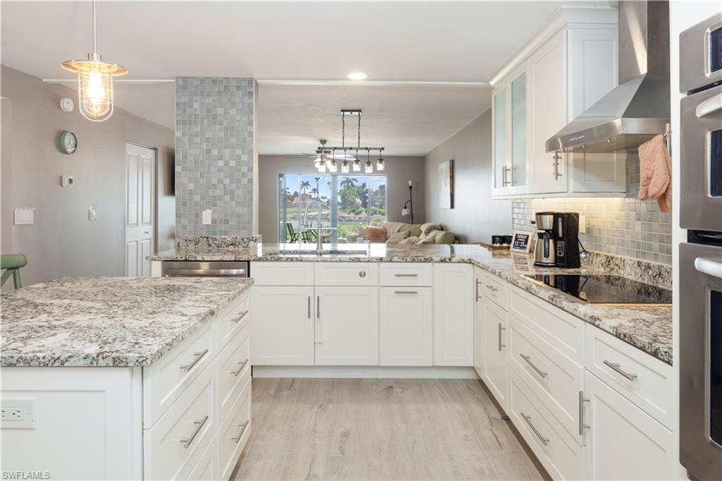 Kitchen featuring decorative light fixtures, stainless steel appliances, kitchen peninsula, white cabinets, and wall chimney range hood