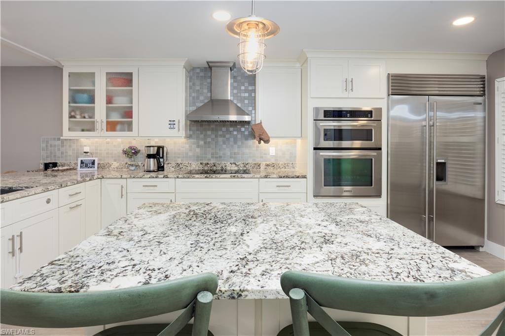 Kitchen featuring backsplash, decorative light fixtures, wall chimney exhaust hood, appliances with stainless steel finishes, and white cabinets