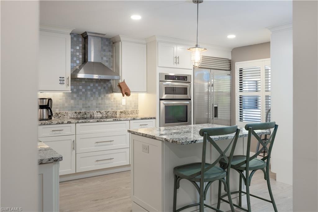 Kitchen featuring decorative light fixtures, wall chimney exhaust hood, stainless steel appliances, white cabinetry, and light wood-type flooring