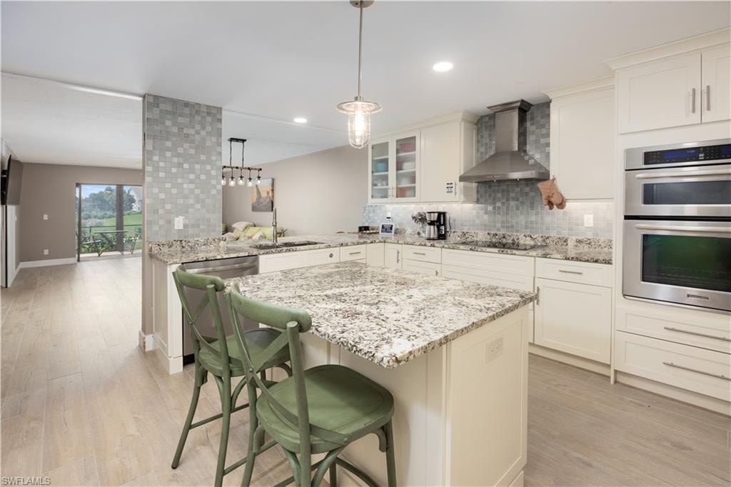 Kitchen with light hardwood / wood-style floors, wall chimney range hood, kitchen peninsula, double oven, and a breakfast bar area