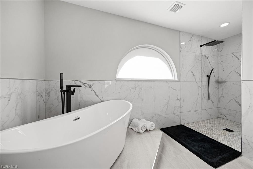 Freestanding soaking tub with a floor-mounted matte black faucet, marble-patterned wall tile, and a walk-in shower with a rainfall showerhead