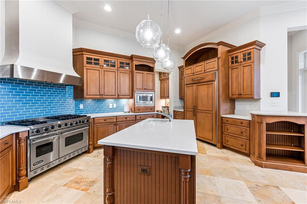 Kitchen featuring wall chimney exhaust hood, hanging light fixtures, built in appliances, a kitchen island with sink, and ornamental molding