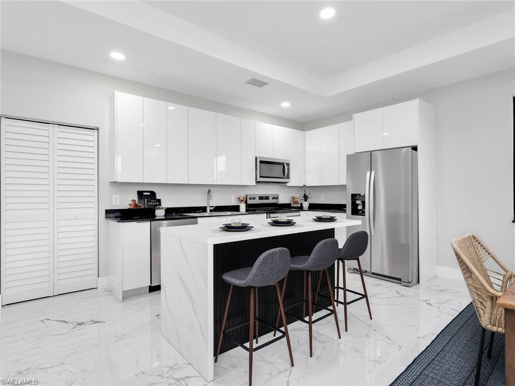 Modern kitchen featuring sleek white cabinetry, black countertops, a waterfall island, stainless steel appliances, and large format polished flooring