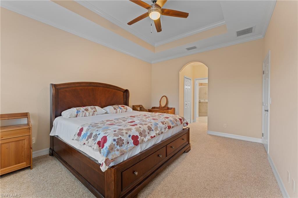 Bedroom with ceiling fan, a tray ceiling, ensuite bath, light colored carpet, and crown molding