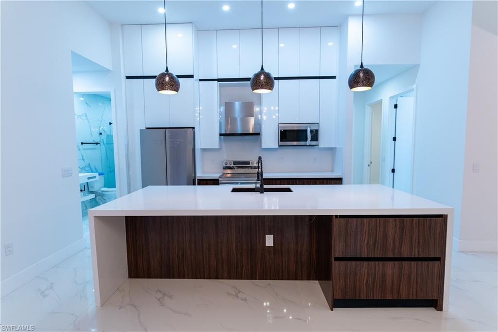 Kitchen featuring appliances with stainless steel finishes, decorative light fixtures, white cabinetry, a kitchen island with sink, and dark brown cabinetry