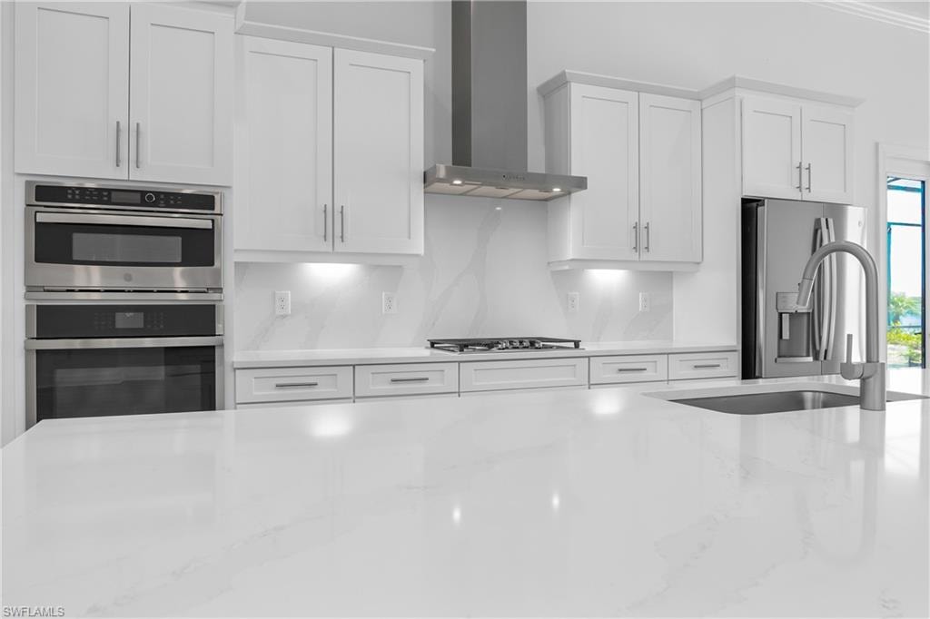 Kitchen featuring white cabinetry, wall chimney range hood, stainless steel appliances, and light stone countertops