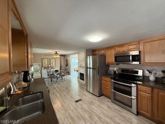 Kitchen with brown cabinetry, dark countertops, appliances with stainless steel finishes, light wood-style floors, and a sink
