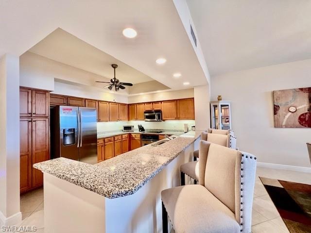 Kitchen with light stone countertops, a peninsula, light tile patterned flooring, stainless steel appliances, and brown cabinets
