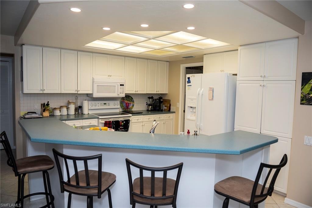 Kitchen features a breakfast bar, tile backsplash, recessed lighting, white appliances and cabinetry, tile floors.