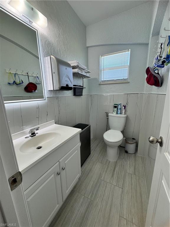Bathroom featuring a wainscoted wall, a textured wall, toilet, vanity, and tile walls