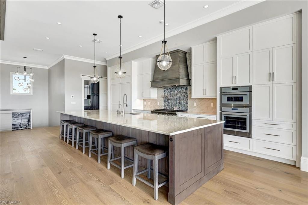 Kitchen with custom exhaust hood, crown molding, white cabinets, light wood-type flooring, and tasteful backsplash