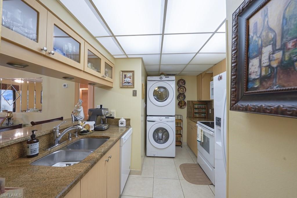 Kitchen featuring stacked washer / dryer, white appliances, light tile patterned floors, glass insert cabinets, and dark stone countertops