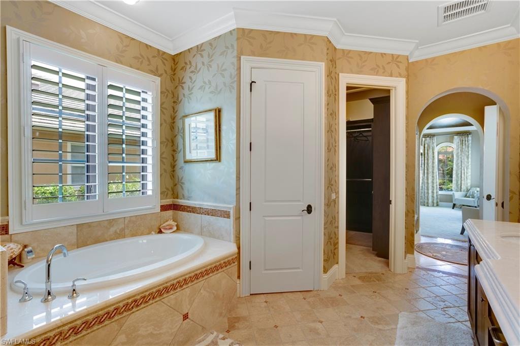 Primary Bathroom with wallpapered walls, ornamental molding, plenty of natural light, a soaking tub, and a large walk-in closet with custom shelving