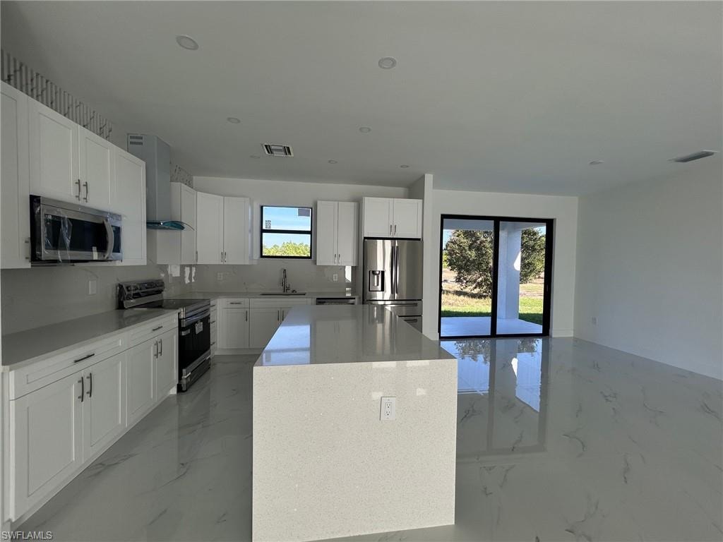 Kitchen featuring appliances with stainless steel finishes, light marble finish flooring, a kitchen island, wall chimney exhaust hood, and recessed lighting