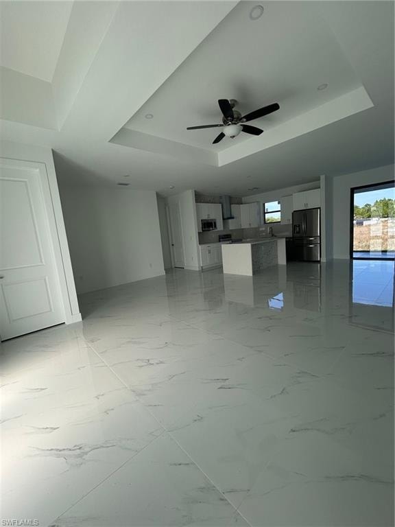 Unfurnished living room featuring a tray ceiling, light marble finish flooring, and a ceiling fan