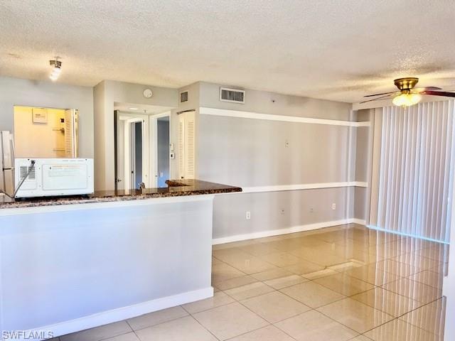 Kitchen with white microwave, tile patterned floors, ceiling fan, a textured ceiling, and dark stone counters