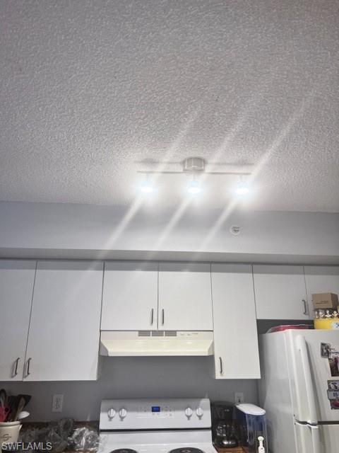 Kitchen view of range hood, refrigerator, stove, white cabinetry, and a textured ceiling