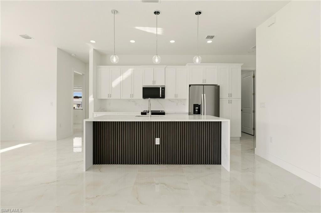 Kitchen with stainless steel fridge, light stone countertops, an island with sink, white cabinets, and pendant lighting
