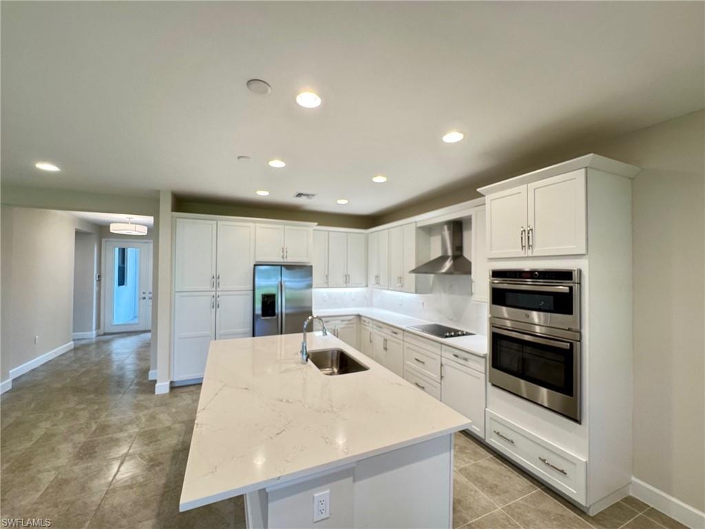 Kitchen with white cabinetry, appliances with stainless steel finishes, recessed lighting, wall chimney exhaust hood, and light stone counters