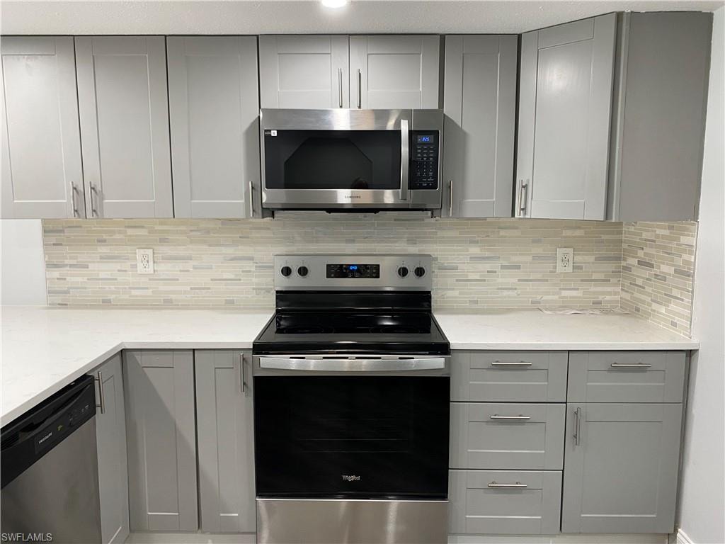Kitchen with gray cabinets, appliances with stainless steel finishes, backsplash, and light stone countertops