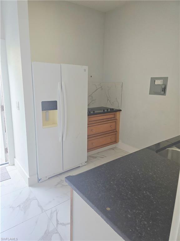 Mother-in-law Kitchen featuring white fridge with ice dispenser, brown cabinetry, light marble finish flooring, and dark stone countertops