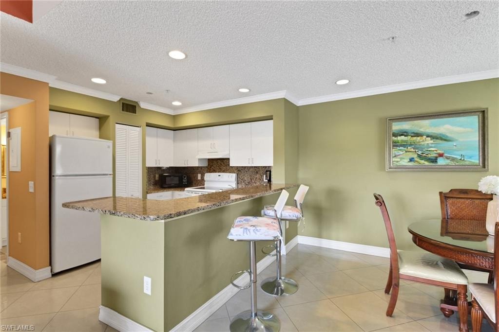 Kitchen featuring white appliances, a kitchen breakfast bar, light tile patterned floors, a textured ceiling, and backsplash