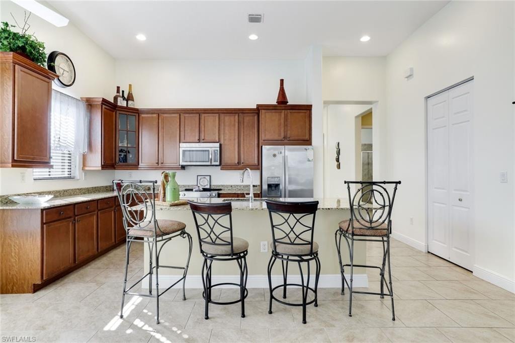 Kitchen with appliances with stainless steel finishes, light stone countertops, glass insert cabinets, a kitchen bar, and brown cabinets