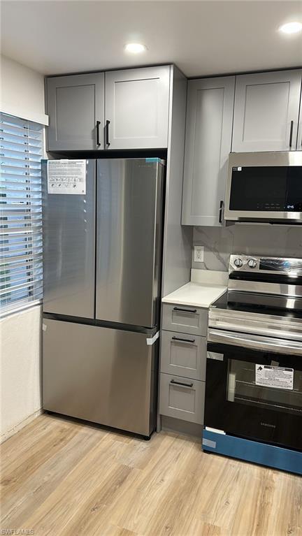 Kitchen featuring gray cabinets, stainless steel appliances, light wood-type flooring, decorative backsplash, and light stone counters
