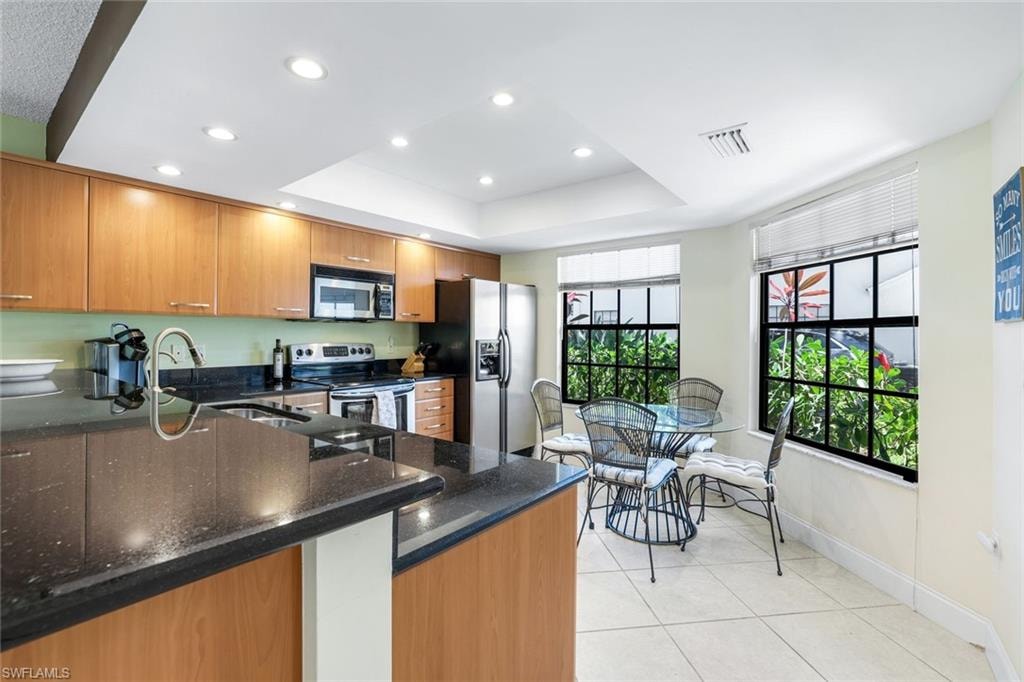Kitchen featuring cherry cabinets, dark stone counters, stainless steel appliances, light tile patterned floors, and a raised ceiling