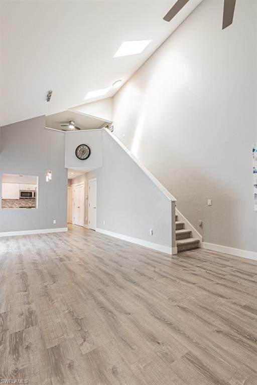 Unfurnished living room featuring a ceiling fan, high vaulted ceiling, light wood finished floors, stairs, and a skylight