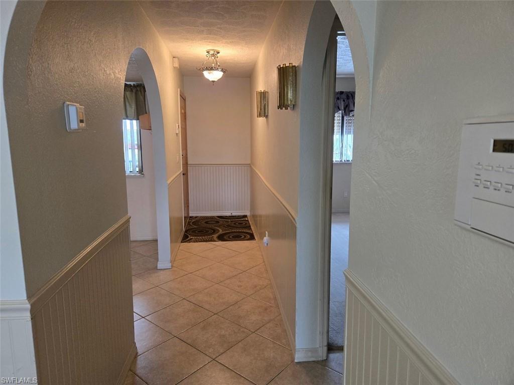 Hallway featuring wainscoting, a textured wall, light tile patterned flooring, arched walkways, and a decorative wall