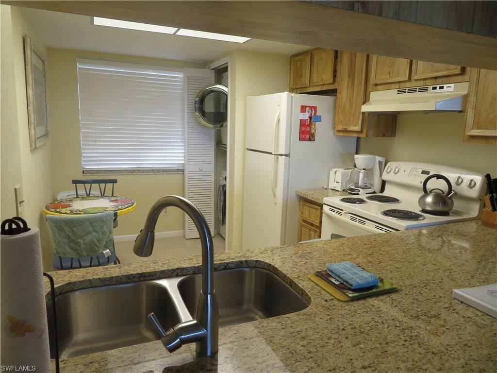 Kitchen with white range with electric stovetop, under cabinet range hood, light stone countertops, and brown cabinets