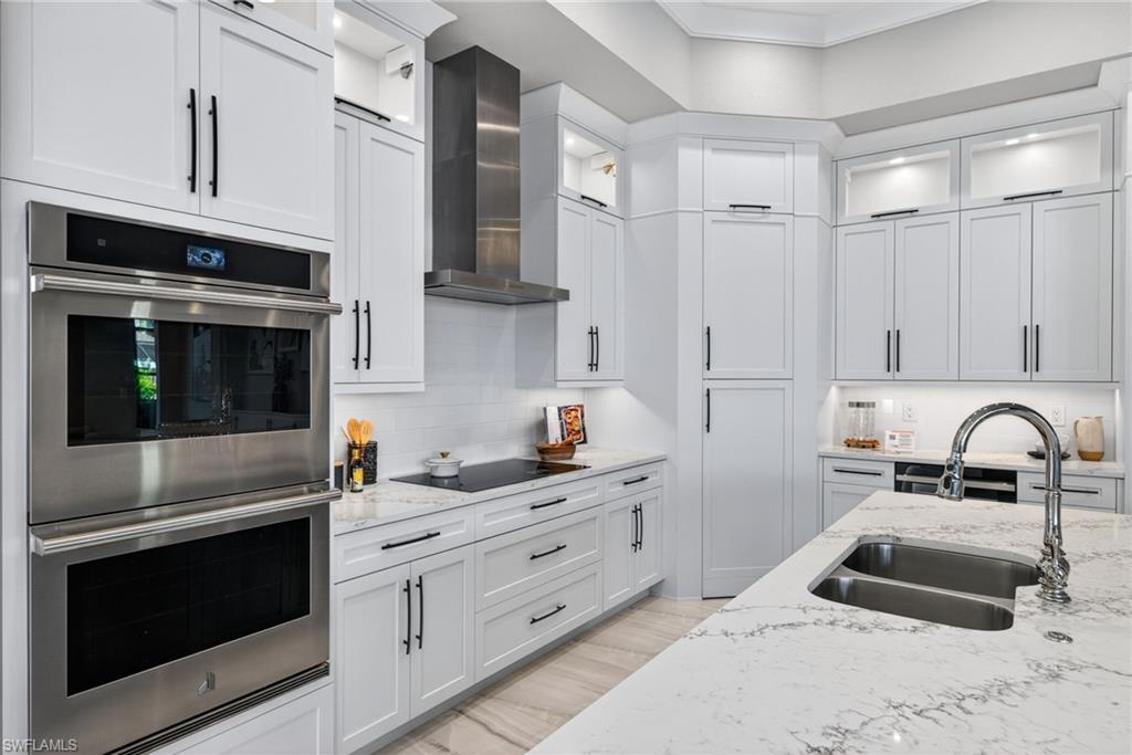 Kitchen featuring stainless steel double oven, glass insert cabinets, light stone countertops, white cabinetry, and wall chimney exhaust hood