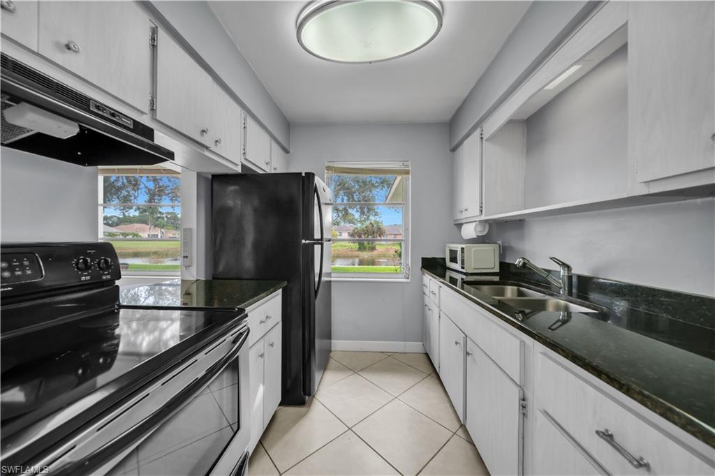 Kitchen with stainless steel electric stove, under cabinet range hood, dark stone countertops, light tile patterned floors, and white microwave
