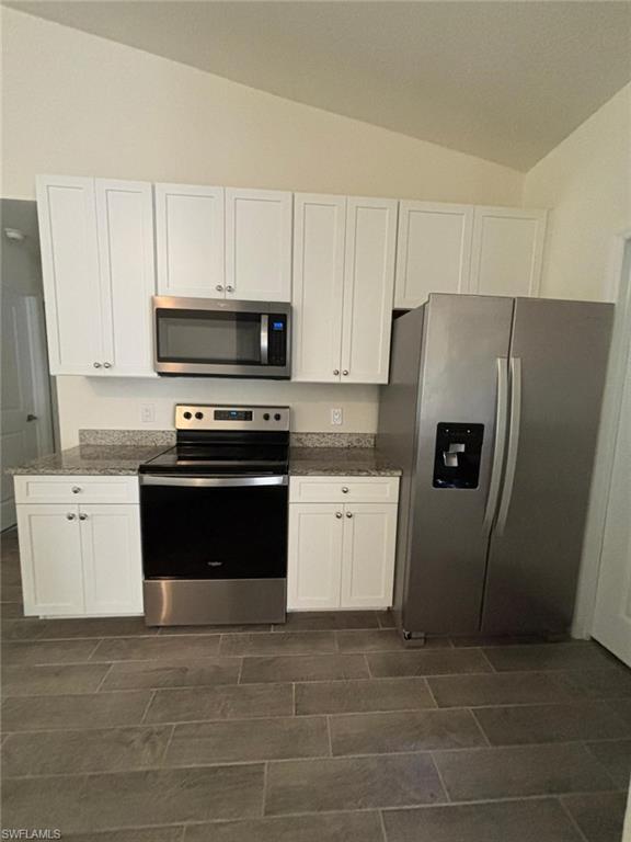 Kitchen featuring white cabinets, stainless steel appliances, lofted ceiling, dark stone counters, and wood finish floors