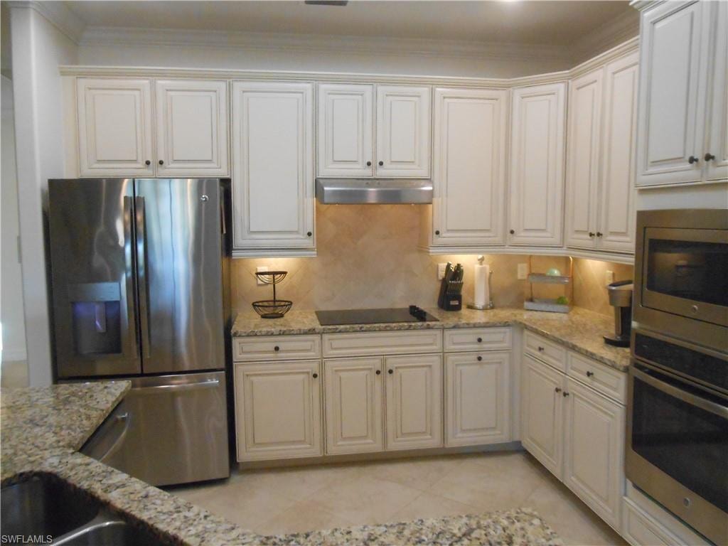 Kitchen with stainless steel appliances, light stone counters, under cabinet range hood, backsplash, and white cabinetry