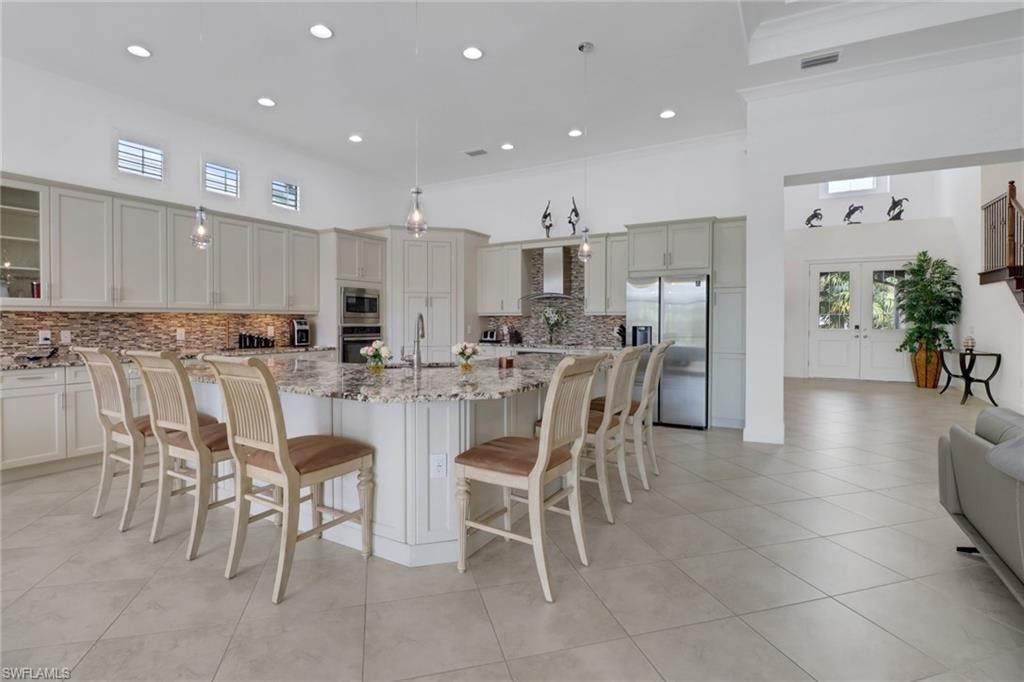 Kitchen featuring wall chimney range hood, appliances with stainless steel finishes, a kitchen island with sink, light stone countertops, and pendant lighting