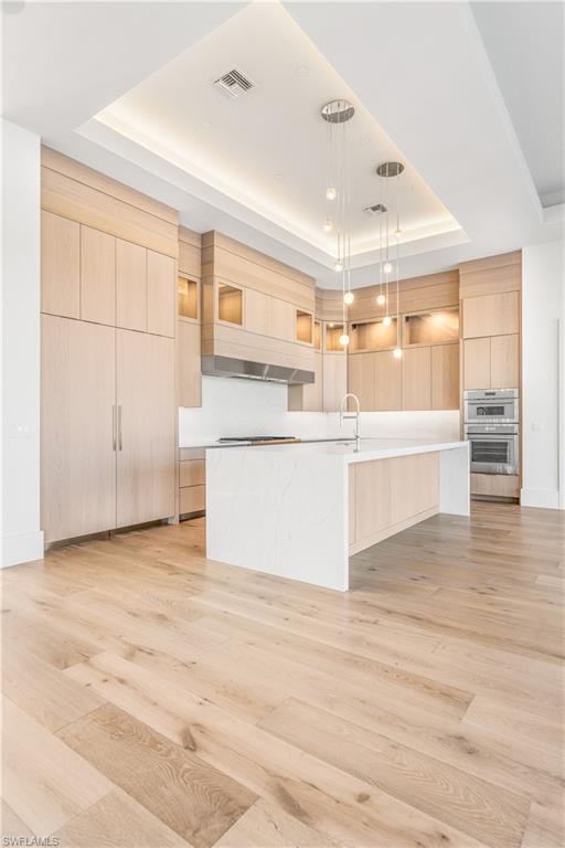 Kitchen with light wood finish cabinetry, a tray ceiling, hanging light fixtures, modern cabinets, and an island with sink