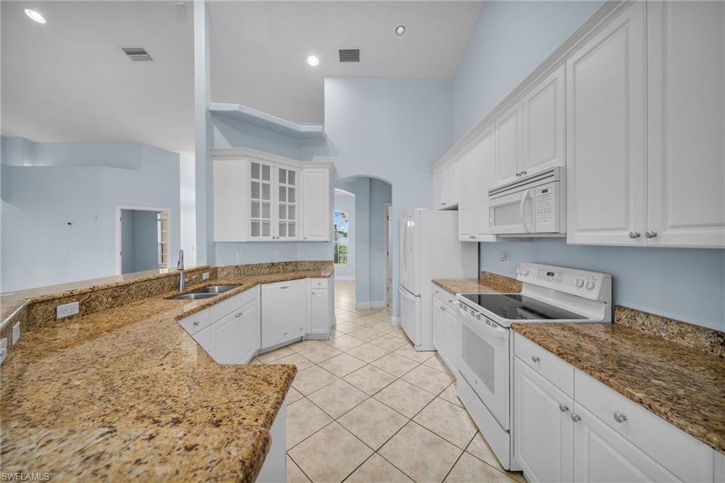 Kitchen with arched walkways, white appliances, glass insert cabinets, white cabinetry, and dark stone counters