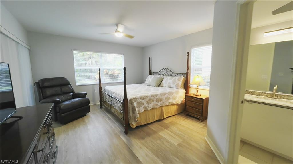 Spacious primary bedroom showcasing a four-poster bed frame, wood-finish flooring, dual windows with natural light, a leather recliner, wood nightstand with attached lighting, and an ensuite bathroom with vanity and mirror visible through the doorway
