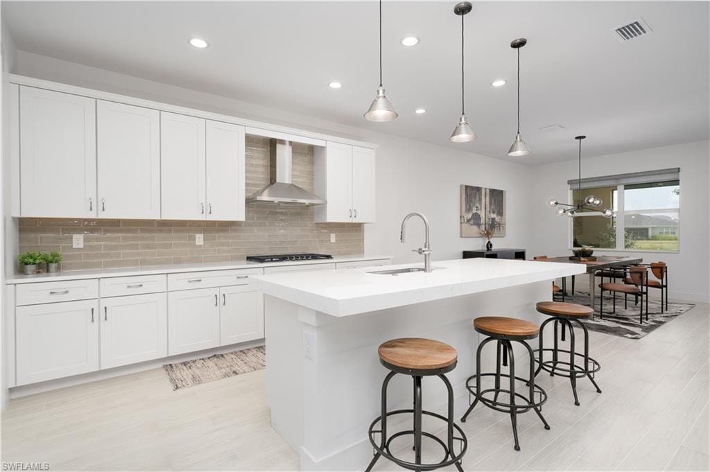 Kitchen with decorative backsplash, decorative light fixtures, an island with sink, wall chimney exhaust hood, and white cabinetry