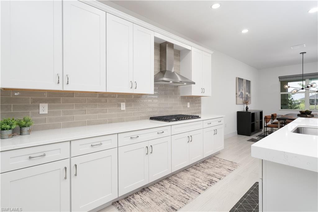 Kitchen featuring wall chimney range hood, light stone countertops, white cabinets, pendant lighting, and backsplash