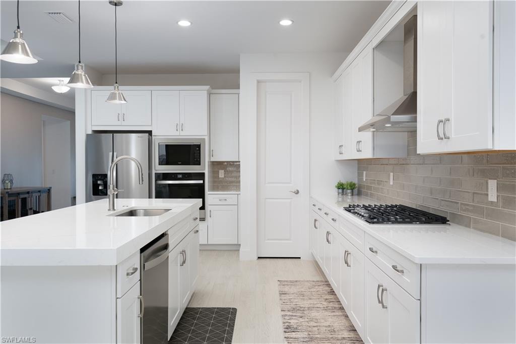 Kitchen with an island with sink, hanging light fixtures, white cabinetry, stainless steel appliances, and light stone counters