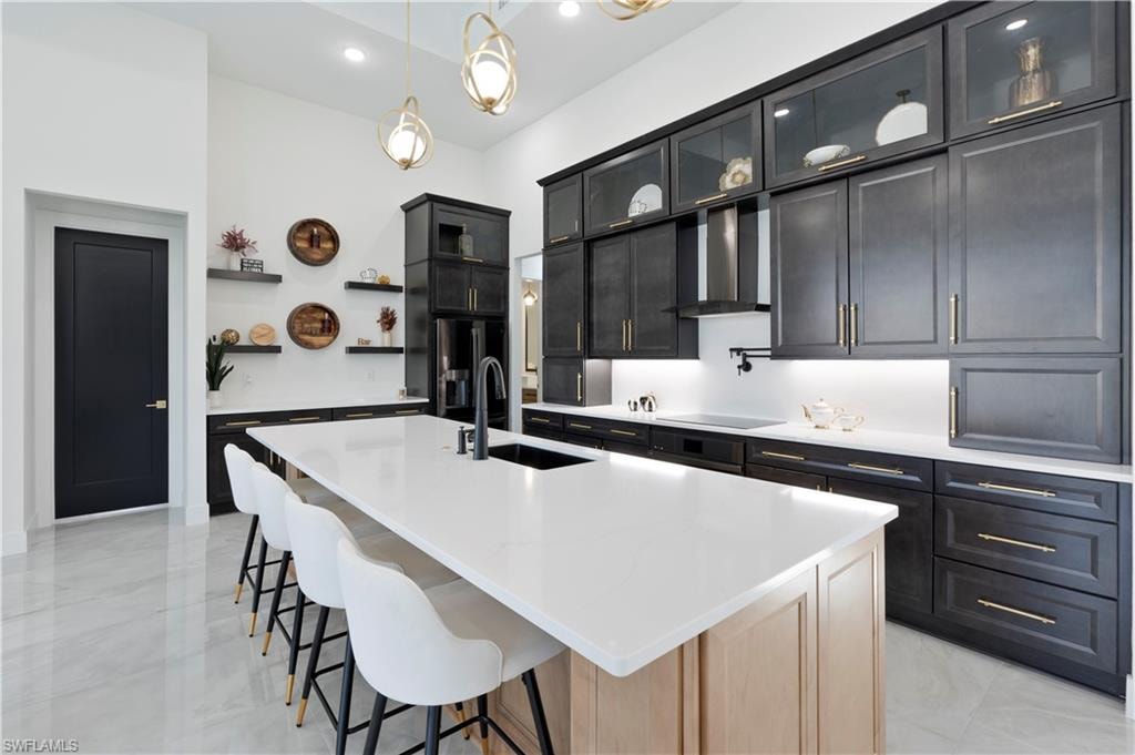 Kitchen with glass insert cabinets, hanging light fixtures, a kitchen island with sink, a breakfast bar, and dark cabinets