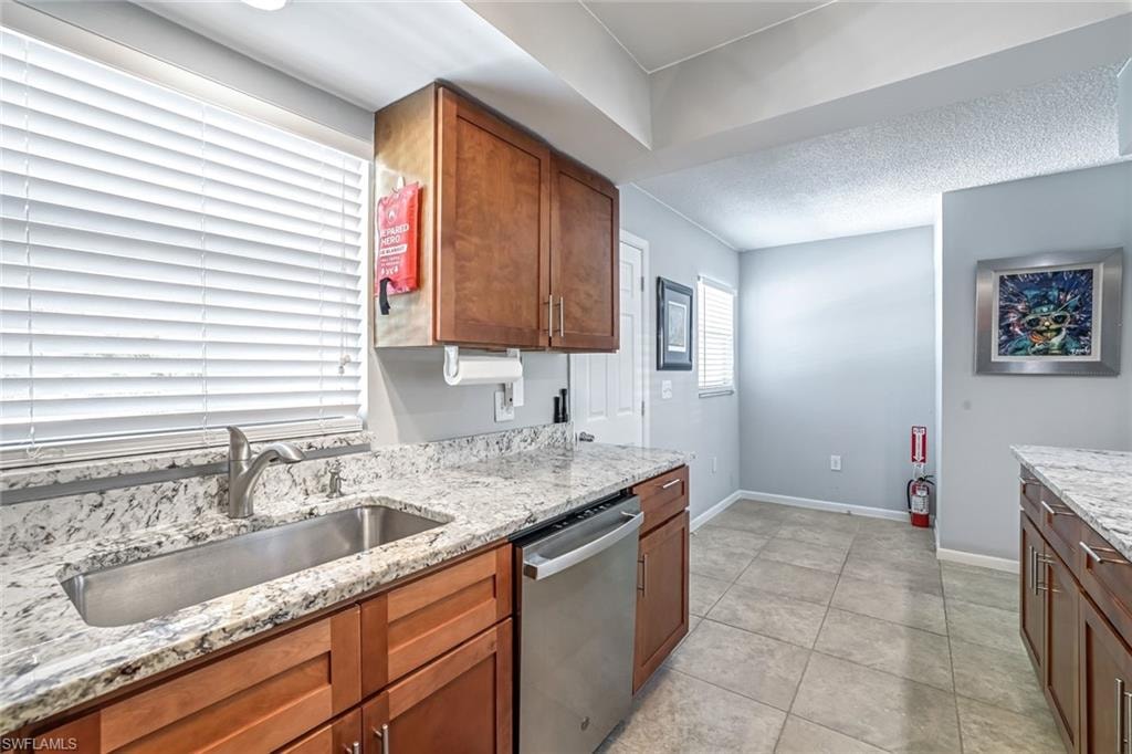Kitchen featuring stainless steel dishwasher, brown cabinetry, light stone counters, a textured ceiling, and light tile patterned floors