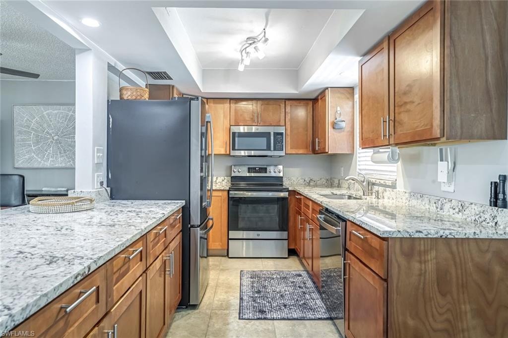 Kitchen featuring stainless steel appliances, light stone counters, brown cabinetry, a tray ceiling, and light tile patterned floors